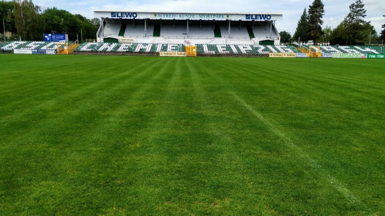 Grüner Rasen vor Tribüne mit grün-weißen-Sitzplätzen.