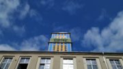 Blau-gelber Turm mit grünem Schild auf saniertem Haus vor blauem Himmel.