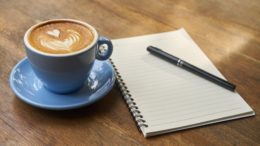 Eine Kaffeetasse und ein Schreibblock mit Stift auf Holzuntergrund.
